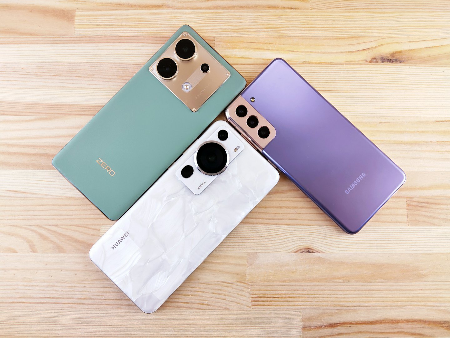 Three modern smartphones rest on a light wooden desk.